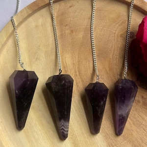 May include: Four amethyst crystal pendulums with silver chains hanging on a wooden plate. The plate is surrounded by rose petals. The text "@SHIFATAGATE" is visible at the bottom of the image.