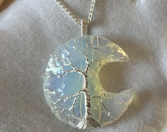 May include: A silver chain necklace with a crescent moon pendant made of opal with a silver wire tree design. The pendant is on a white surface.