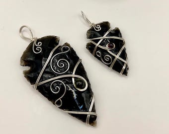 May include: Two black arrowhead pendants with silver wire detailing, one larger and one smaller, are displayed. The pendants are set against a white background, with a dark crystal formation in the background. The text "@SHIFATAGATE" is visible.