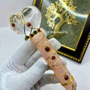 Op de afbeelding: Een kristallen toverstaf met een heldere hartvormige top en een roze rozenkwarts lichaam. De toverstaf is versierd met goudkleurige draad en rode en groene edelstenen. De toverstaf wordt vastgehouden door een gehandschoende hand. De tekst "@Shifatagate" is zichtbaar.
