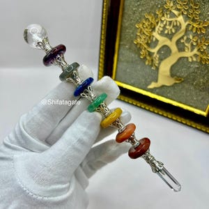 May include: A silver wand with a clear crystal ball at the top and a clear crystal point at the bottom. The wand features seven colorful gemstone discs in shades of red, orange, yellow, green, blue, and purple. The wand is held in a white gloved hand.