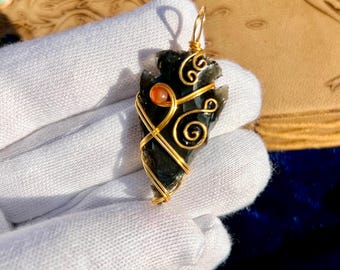 May include: A black arrowhead pendant wrapped in gold wire, with a small orange bead accent. The pendant is held against a blue background, with a leather-bound book with a tree design in the background.
