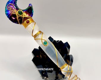 May include: A handcrafted crystal wand with a crescent moon-shaped top. The wand features a clear crystal shaft wrapped in gold wire, adorned with green and red gemstones. The moon is iridescent with gold wire accents.