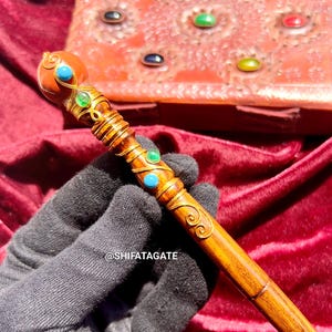 May include: A handcrafted wooden wand with gold accents and turquoise and green gemstones. The wand has a rounded top and a pointed tip. The wand is held against a red velvet background. The wand is approximately 12 inches long.
