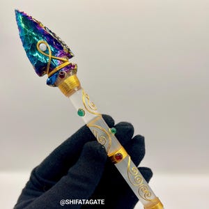 Puede incluir: Una varita decorativa con una punta de flecha iridiscente y colorida. La varita presenta detalles de alambre dorado, vidrio transparente y pequeñas piedras preciosas. El texto "@SHIFATAGATE" es visible.