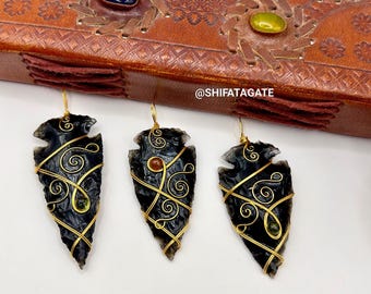 May include: Three black arrowhead earrings with gold wire detailing and small gemstone accents. The earrings are displayed against a white background, with a brown leather journal in the background.