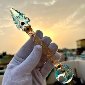 May include: A decorative dagger with a clear handle and a blue-tinted blade. The blade has a unique arrowhead shape with a curved design at the base. The handle is wrapped with gold-coloured wire and adorned with red and green accents. The dagger is held in a white glove.