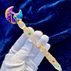 May include: A handcrafted crystal wand with a crescent moon-shaped top. The wand features a clear crystal shaft wrapped in gold wire, adorned with green and red gemstones. The moon is iridescent with purple, blue, and gold hues.