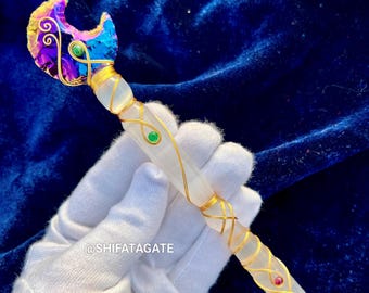 May include: A handcrafted crystal wand with a crescent moon-shaped top. The wand features a clear crystal shaft wrapped in gold wire, adorned with green and red gemstones. The moon is iridescent with purple, blue, and gold hues.