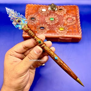 May include: A handcrafted wooden pen with a clear, blue crystal point and gold wire accents. The pen features a brown wooden body with gold filigree and turquoise and green gemstone embellishments. The pen has a brass tip.