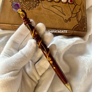 Puede incluir: Un bolígrafo de madera con un cristal de amatista púrpura en la parte superior, detalles dorados y un cuerpo de madera marrón. El bolígrafo se sostiene en una mano enguantada. El texto "@SHIFATAGATE" es visible en el fondo.