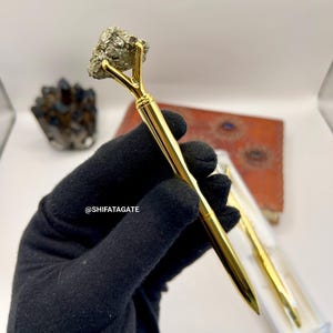 May include: A gold-colored pen with a pyrite stone at the top. The pen is held by a gloved hand. The pen is in a clear plastic case. The text "@SHIFATAGATE" is visible.