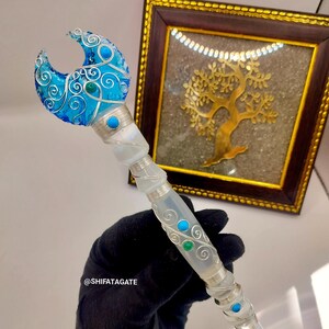 Peut inclure: Une baguette artisanale avec un dessus en forme de croissant de lune bleu, orné de fil d'argent et d'accents turquoise. Le manche transparent est orné de fil d'argent et d'embellissements turquoise et verts. La baguette est tenue par une main gantée.