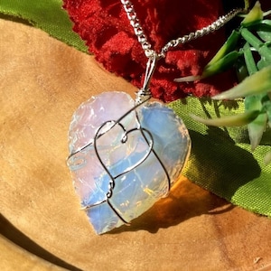 May include: A silver chain necklace with a heart-shaped opal pendant wrapped in silver wire. The pendant is iridescent and has a light blue and white colour.
