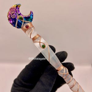 May include: A handcrafted crystal wand with a crescent moon-shaped top. The moon is adorned with colorful swirls and gems. The wand is wrapped in copper wire and features red and green gemstones. The wand is held by a gloved hand.