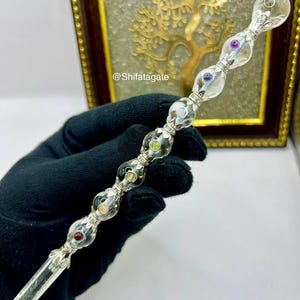 May include: A clear crystal wand with a silver-colored metal handle and colorful gemstone accents. The wand features a faceted crystal tip and a series of clear, rounded glass beads with embedded gems. The background includes a framed artwork with a golden tree design.