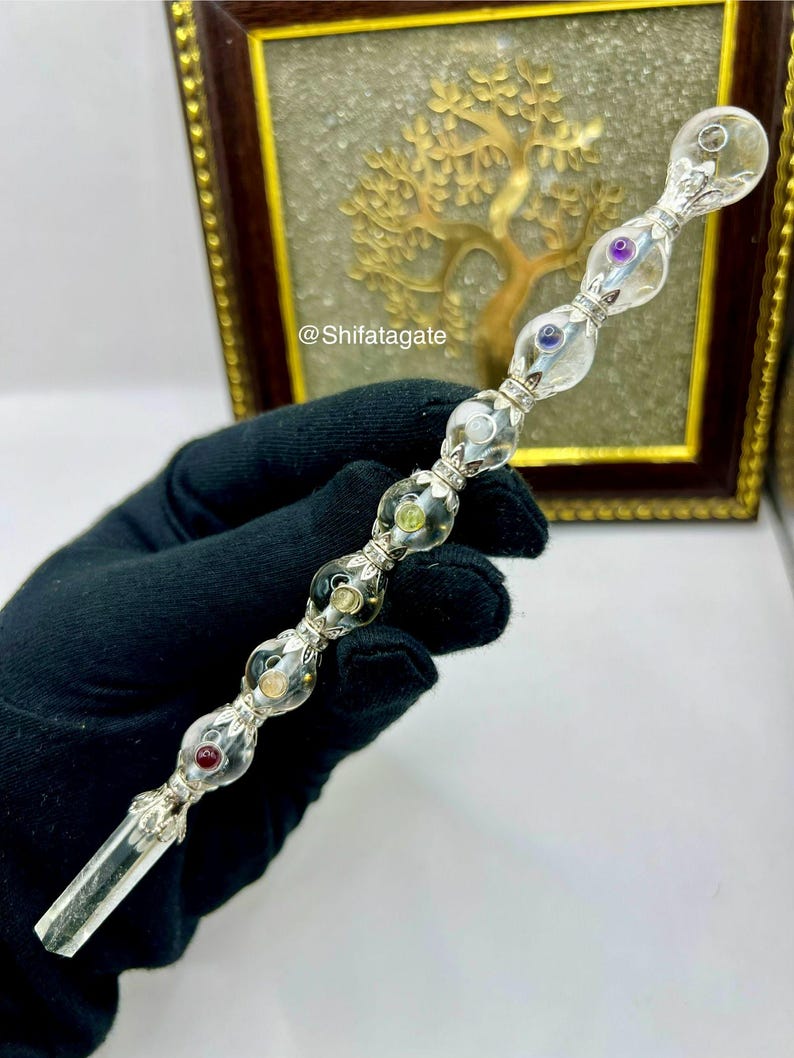 May include: A clear crystal wand with a silver-colored metal handle and colorful gemstone accents. The wand features a faceted crystal tip and a series of clear, rounded glass beads with embedded gems. The background includes a framed artwork with a golden tree design.