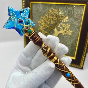 May include: A handcrafted wand with a blue star-shaped crystal top. The wand has a dark wood handle wrapped with gold wire and gemstones. The wand is held in a white gloved hand.