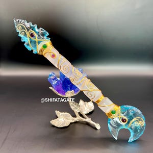 Puede incluir: Una varita de cristal decorativa con un diseño transparente, azul y dorado. La varita presenta una hoja de cristal transparente, un mango blanco y dorado y está adornada con piedras preciosas de colores. La varita se exhibe con un soporte plateado en forma de hoja.