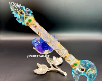 May include: A decorative crystal wand with a clear, blue, and gold design. The wand features a clear crystal blade, a white and gold handle, and is adorned with colorful gemstones. The wand is displayed with a silver leaf-shaped stand.