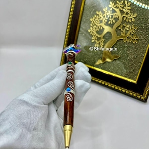 May include: A handcrafted pen with a dark brown wooden barrel, adorned with silver accents and turquoise and green embellishments. The pen's top features a decorative design with iridescent details. The pen has a gold-colored tip.