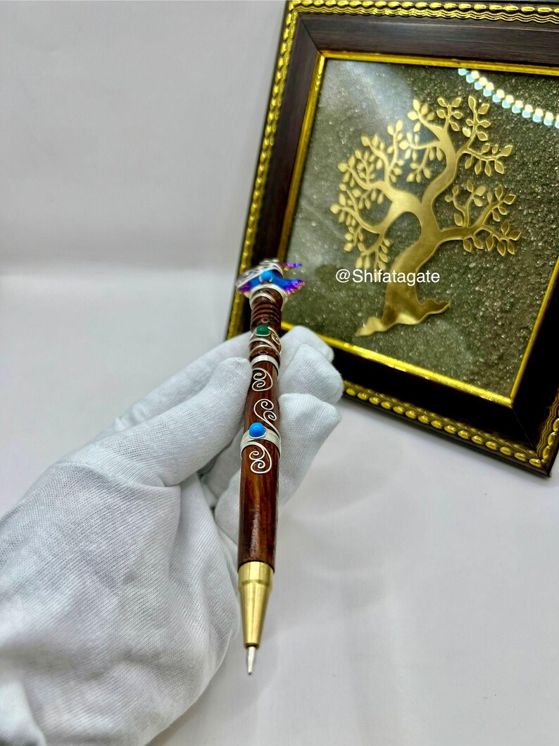 May include: A handcrafted pen with a dark brown wooden barrel, adorned with silver accents and turquoise and green embellishments. The pen's top features a decorative design with iridescent details. The pen has a gold-colored tip.