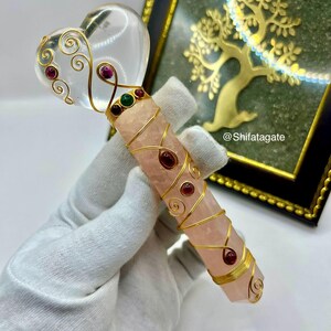 Op de afbeelding: Een handgemaakte kristallen toverstaf met een heldere hartvormige top en een roze rozenkwarts lichaam. De toverstaf is versierd met gouden draad en rode en groene edelstenen. Op de achtergrond een gouden boom in een lijst.