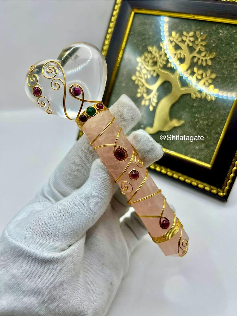 Op de afbeelding: Een handgemaakte kristallen toverstaf met een heldere hartvormige top en een roze rozenkwarts lichaam. De toverstaf is versierd met gouden draad en rode en groene edelstenen. Op de achtergrond een gouden boom in een lijst.