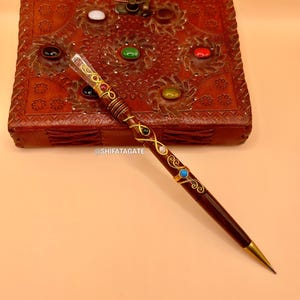 May include: A dark brown pen with gold accents and colorful gemstones. The pen is next to a brown leather box with carved floral designs and inlaid gemstones. The pen has a gold tip and is decorated with swirls.