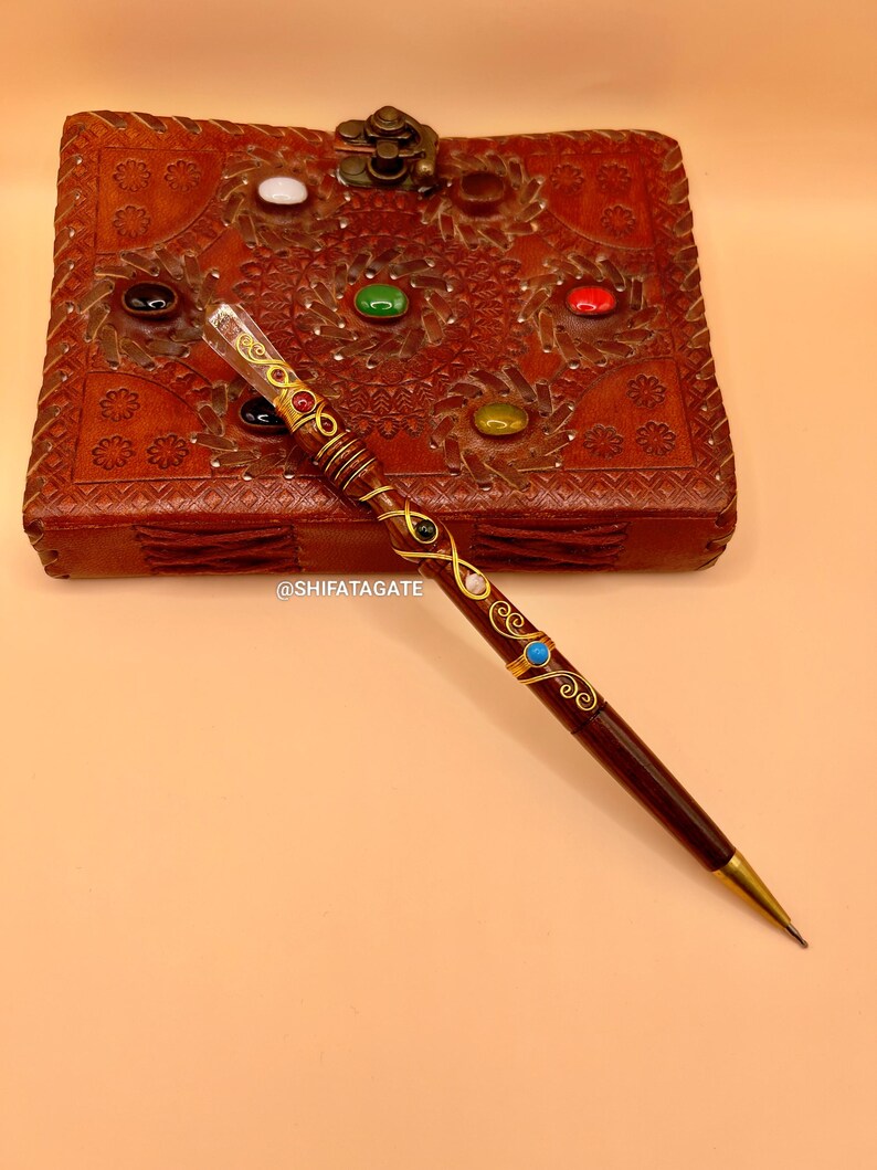 May include: A dark brown pen with gold accents and colorful gemstones. The pen is next to a brown leather box with carved floral designs and inlaid gemstones. The pen has a gold tip and is decorated with swirls.