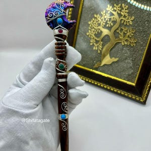 May include: A handcrafted pen with a dark brown wooden barrel and a decorative top. The top features a crescent moon design with iridescent colors, turquoise, and red accents. Silver wire detailing and turquoise accents adorn the barrel. The pen is held by a gloved hand.