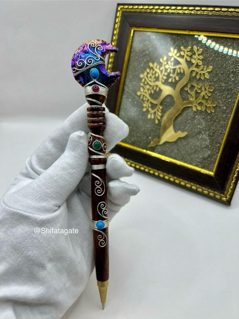 May include: A handcrafted pen with a dark brown wooden barrel and a decorative top. The top features a crescent moon design with iridescent colors, turquoise, and red accents. Silver wire detailing and turquoise accents adorn the barrel. The pen is held by a gloved hand.