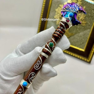 May include: A handcrafted wooden wand with a carved bear head, adorned with silver wire, colorful gemstones, and a swirling design. The bear head is a mix of blue, purple, and iridescent colors. The wand's handle is dark brown with silver wire accents.