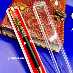 K&ouml;nnte beinhalten: Ein schwarzer und goldener Stift in einer wei&szlig;en Box mit rotem Innenraum, neben einer transparenten Kunststoffh&uuml;lle. Der Stift hat einen dekorativen Schaft mit goldenen Akzenten. Der Stift liegt auf einem braunen Lederjournal mit farbenfrohen Verzierungen. Der Text "@SHIFATAGATE" ist sichtbar.