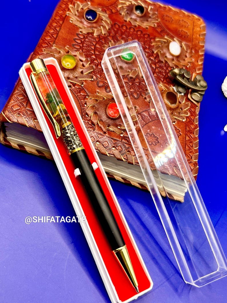 K&ouml;nnte beinhalten: Ein schwarzer und goldener Stift in einer wei&szlig;en Box mit rotem Innenraum, neben einer transparenten Kunststoffh&uuml;lle. Der Stift hat einen dekorativen Schaft mit goldenen Akzenten. Der Stift liegt auf einem braunen Lederjournal mit farbenfrohen Verzierungen. Der Text "@SHIFATAGATE" ist sichtbar.