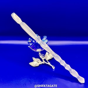 May include: A clear, elongated crystal wand with a wavy design, resting on a silver-colored metal rose with blue and purple crystal accents. The background is a solid, deep blue. The text "@SHIFATAGATE" is at the bottom.