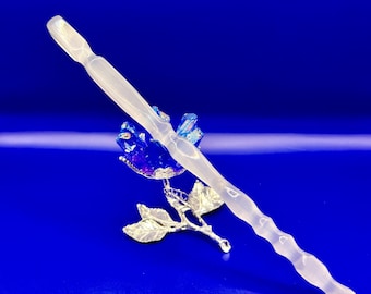 May include: A clear, elongated crystal wand with a wavy design, resting on a silver-colored metal rose with blue and purple crystal accents. The background is a solid, deep blue. The text "@SHIFATAGATE" is at the bottom.