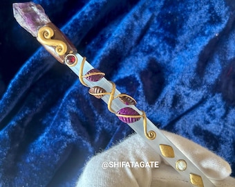 May include: A white crystal wand with gold wire wrapped around it. The wand is decorated with purple and brown accents, including a purple crystal at the top and a small pearl bead. The wand is held in a white gloved hand. The text "@SHIFATAGATE" is visible on the wand.