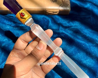 May include: A white selenite wand with a purple amethyst crystal tip and a gold-coloured metal band with a red gemstone. The wand is held in a hand against a blue background. @shifatagate