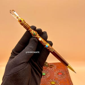 May include: A decorative pen with a clear crystal tip and a dark brown wooden body. The pen is adorned with gold-colored metal accents and colorful gemstones. The pen is held by a gloved hand. The text "@SHIFATAGATE" is visible.