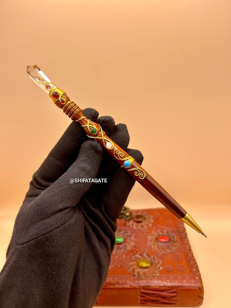 May include: A decorative pen with a clear crystal tip and a dark brown wooden body. The pen is adorned with gold-colored metal accents and colorful gemstones. The pen is held by a gloved hand. The text "@SHIFATAGATE" is visible.