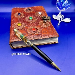 K&ouml;nnte beinhalten: Ein schwarzer und goldener Stift mit transparentem Schaft liegt auf einer blauen Oberfl&auml;che neben einem braunen Lederjournal mit bunten Edelsteinen und einer dekorativen blauen Blume. Das Journal hat ein aufwendiges gepr&auml;gtes Design.