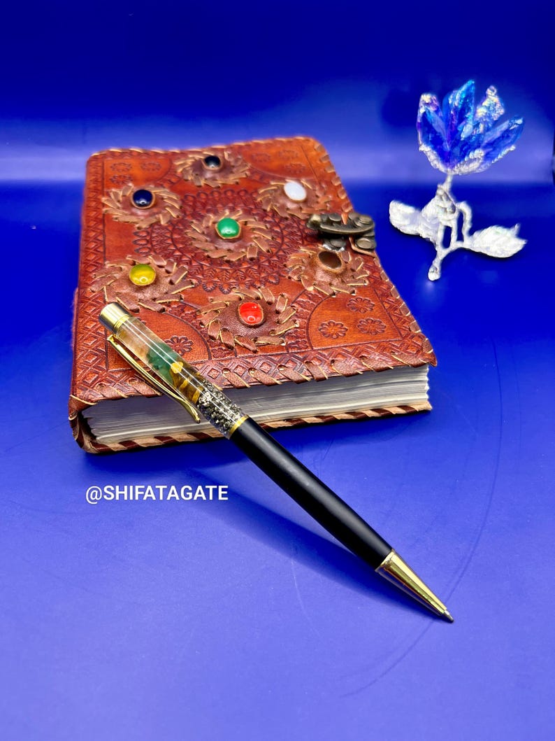 K&ouml;nnte beinhalten: Ein schwarzer und goldener Stift mit transparentem Schaft liegt auf einer blauen Oberfl&auml;che neben einem braunen Lederjournal mit bunten Edelsteinen und einer dekorativen blauen Blume. Das Journal hat ein aufwendiges gepr&auml;gtes Design.