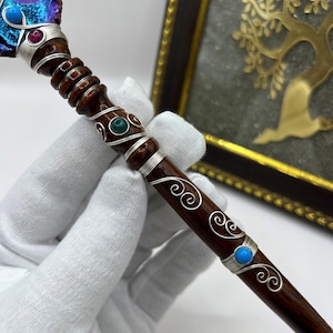 May include: A handcrafted wooden wand with a dark brown finish, adorned with silver wire, and colorful gemstones. The wand's top features a blue and purple crystal, while the shaft is embellished with turquoise, red, and green stones. The wand is held by a gloved hand.