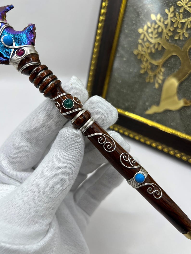 May include: A handcrafted wooden wand with a dark brown finish, adorned with silver wire, and colorful gemstones. The wand's top features a blue and purple crystal, while the shaft is embellished with turquoise, red, and green stones. The wand is held by a gloved hand.