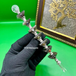 May include: A silver-toned wand with a clear crystal at the base and a clear orb at the top. The wand features stacked, geometric, gray-brown stones with colorful gemstones and ornate silver accents. The background is a framed gold tree on a textured surface.