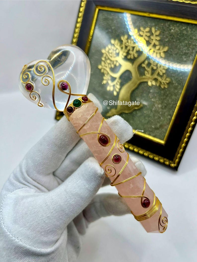 Op de afbeelding: Een kristallen toverstaf met een heldere hartvormige top en een roze rozenkwarts lichaam. De toverstaf is versierd met goudkleurige draad en rode en groene edelstenen. De toverstaf wordt vastgehouden door een gehandschoende hand. De tekst "@Shifatagate" is zichtbaar.