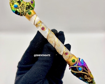 May include: A handcrafted wand with a white crystal shaft, adorned with gold wire, and colorful gemstones. The wand features a crescent moon and arrowhead design, with iridescent and jewel-toned accents. The text "@SHIFATAGATE" is visible.