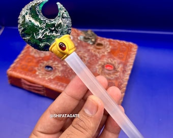 May include: A fantasy-themed staff with a green, crescent-shaped top. The staff has a gold-colored band with a red gem and a clear, cylindrical handle. The background is a blue surface.