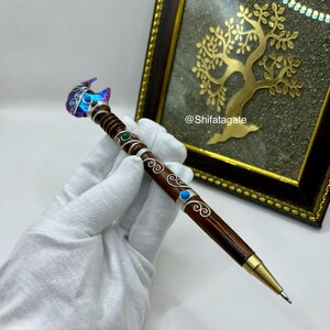 May include: A brown wooden pen featuring silver wire filigree, small green and blue inlaid stones, and a metallic gold tip. The top of the pen is adorned with a faceted, iridescent blue and purple crystal. The text @Shifatagate is visible.